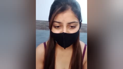 Media: Video of a young South Asian woman with long brown hair, wearing a black face mask, maroon tank top, and a plain background.