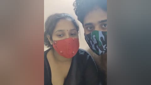 Media: Video of two South Asian individuals with medium skin tones, one wearing a red mask and the other a camouflage mask, both with short hair, in a blurred indoor setting.