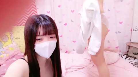 Media: A video of a young Asian woman with long black hair, wearing a white face mask, sitting on a pink bed with hearts patterned curtains. A partially visible person, also masked, stands behind her.