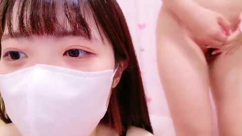 Media: Video of an Asian woman with long black hair and brown eyes, wearing a white face mask, positioned close to a nude person's exposed genitalia.