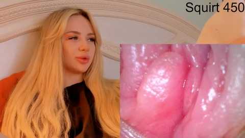 Video of a young blonde woman with long hair, wearing a black top, sitting on a bed with a beige headboard. Close-up of her pink, moist, vaginal tissue is superimposed on the image.