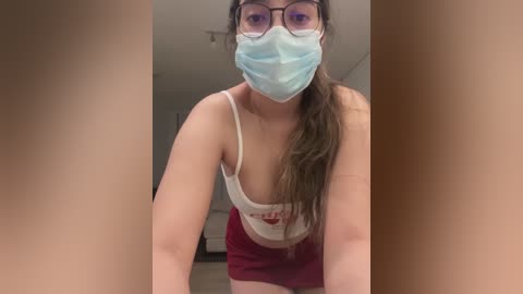 A video of a young woman with long brown hair, wearing a white tank top and red shorts, a blue mask, and glasses, standing indoors.