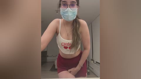 Media: A video of a young woman in a white \"Chili's\" crop top and red shorts, wearing a mask, standing indoors with a blurred background.