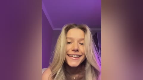 Media: A video of a smiling, topless young woman with long, blonde hair, standing indoors under purple lighting. She has a light complexion and is wearing a necklace.