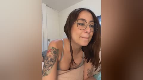 Media: Video of a light-skinned woman with long brown hair and glasses, wearing a beige tank top, showing off colorful tattoos on her arms, in a dimly lit room with white walls and a door.