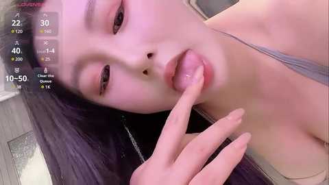 Media: Video of a young East Asian woman with long black hair, wearing a gray tank top, lying on a wooden floor, licking her index finger, with a digital thermometer overlay showing 40.5\u00b0C.