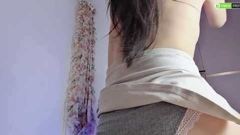 Media: A video of a woman's lower back and buttocks in a light gray, lace-trimmed dress, standing in a room with a partially visible white wall and a purple, textured hanging object.