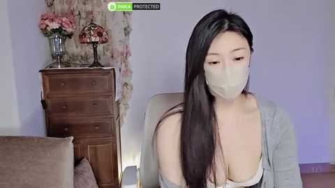 Media: A video of an Asian woman with long black hair, wearing a mask, gray cardigan, and a revealing white top, seated indoors with floral-patterned curtains and a wooden dresser in the background.