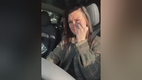 Media: Video of a woman with shoulder-length brown hair, wearing a plaid shirt, sitting in a car with a distressed expression, covering her mouth.