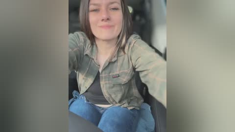 Media: Video of a smiling, light-skinned woman with shoulder-length brown hair, wearing a green plaid jacket over a black shirt and blue jeans, seated in a car.