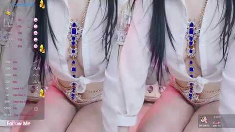 Media: A video of a woman with long black hair, wearing a sheer white blouse and a beaded blue necklace, sitting on a bed with a blurred background.