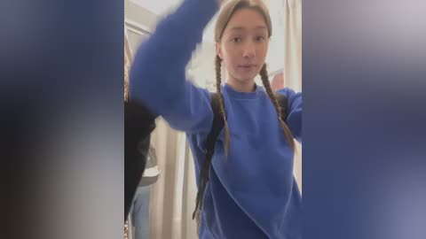 Media: Video of a young Asian girl with long, blonde pigtails, wearing a blue sweater, standing in a dimly lit room with beige curtains and a black backpack.