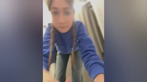 Media: Video of a young woman with light skin and long, brown braided hair, wearing a blue sweater and jeans, bending down, looking at a smartphone, in a bathroom with beige walls and a shower curtain.