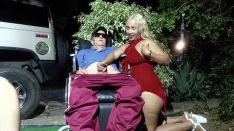 Media: Video of an elderly man in a wheelchair wearing a blue shirt and red pants, with a blonde woman in a red dress, both outdoors at night.