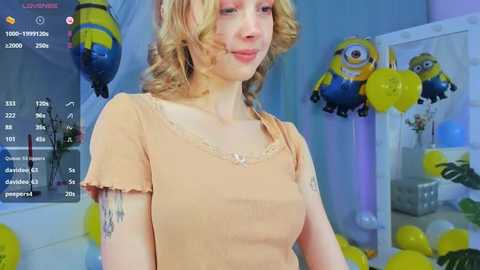 Media: Video of a fair-skinned, blonde woman with curly hair in a beige dress, surrounded by yellow balloons and Minions decorations.