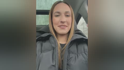 Media: Video of a smiling, fair-skinned woman with long blonde hair, wearing a dark puffer jacket, sitting in a car.