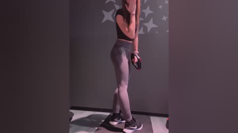 Media: Video of a woman in a black sports bra and grey leggings, holding a black dumbbell, standing on a balance board in a dimly lit room with grey walls and star decals.