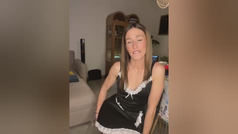 Media: Video of a young woman in a black satin maid outfit with white lace, sitting on a couch, eyes closed, in a dimly lit living room.