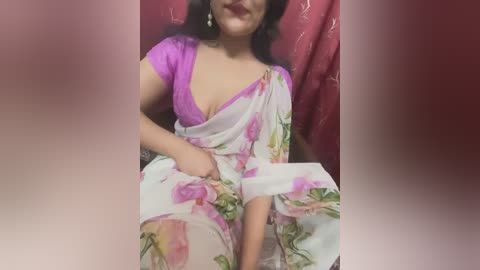 Media: A video of a South Asian woman in a floral sari with a deep purple blouse, sitting against a red patterned background. She has long, dark hair and is smiling.