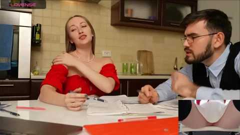 Media: Video of a woman in a red dress and man in glasses, both seated at a kitchen table, discussing papers. Background includes beige tiled walls, dark cabinets, and a stainless steel refrigerator.