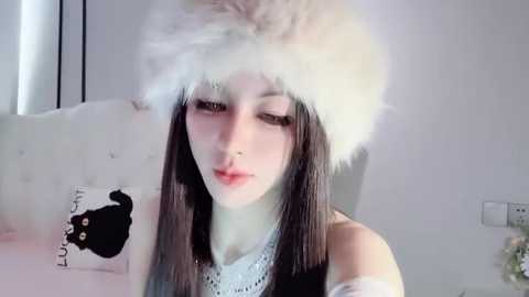 Media: Video of a young woman with fair skin, long dark hair, and a white fur hat, wearing a white sweater and sitting on a bed with a black bear print pillow.