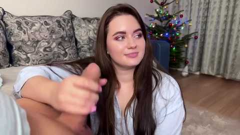 Media: Video of a young, fair-skinned woman with long brown hair, wearing a white robe, sitting on a sofa with patterned pillows. She holds a penis in her hand. Christmas tree with colorful lights in the background.