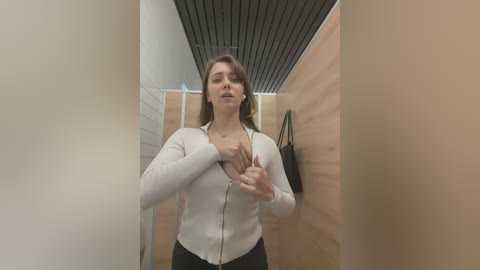 Media: Video of a woman with medium skin tone, light brown hair, wearing a white zip-up cardigan and black pants, standing in a narrow, wooden-walled bathroom with a black ceiling. She covers her chest with her hands.