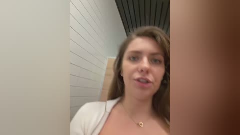 Media: Video of a young Caucasian woman with light skin and brown hair, wearing a white cardigan, standing in a white-tiled hallway with wooden ceiling panels. She has a neutral expression.