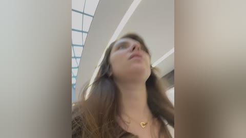 Media: A video of a young woman with long brown hair, wearing a heart-shaped necklace, standing in a modern building with a glass ceiling.