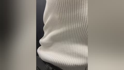 Media: Video of a close-up of a person's torso wearing a white, ribbed, textured sweater and black leather pants. The sweater is slightly wrinkled, emphasizing its texture. The background is blurred, with a dark grey tone.