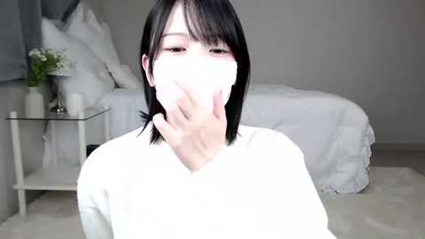 Media: Video of a young East Asian woman with short black hair, wearing a white mask, sitting on a white rug in a minimalist bedroom with a white bed and nightstand.