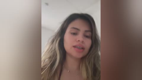 Media: Video of a young woman with long, wavy, dark blonde hair, light skin, and a neutral expression. The background is blurred, likely indoors with a plain, off-white wall.