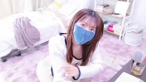 Media: Video of an East Asian woman with long brown hair and a blue face mask, sitting on a pink shag rug in a tidy, white bedroom with a bed, nightstand, and chair.