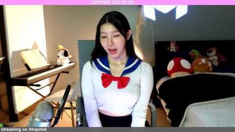 Media: A video of an Asian woman with straight black hair, wearing a white sailor school uniform with a red bow, singing into a microphone in a brightly lit room.