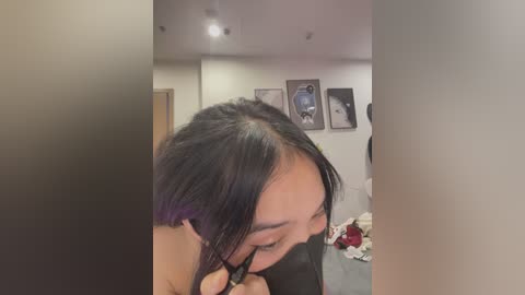 Media: Video of a young Asian woman with long, dark hair, braiding it with a black hair tie. She's indoors in a dimly lit room with white walls, a bed, and framed art pieces in the background.