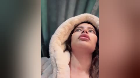 Media: Video of a young woman with fair skin, brown hair, and light pink lipstick, wearing a plush white hooded robe, looking upward with a serene expression against a muted green curtain background.