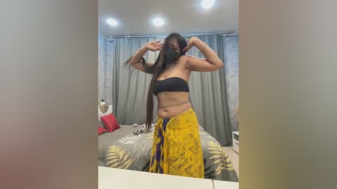 Media: Video of a woman with long black hair, wearing a black strapless top and yellow sarong, posing with arms raised in a bedroom with gray curtains and a bed.