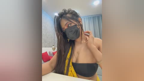 Media: Video of a South Asian woman with long, dark hair and medium skin tone, wearing a black face mask, black bra, and yellow sari, seated indoors, with a modern, minimalist background.