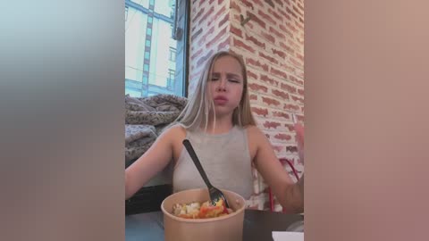 Video of a blonde woman with long hair, wearing a sleeveless top, eating soup from a bowl with a spoon, in a cozy caf\u00e9 with brick walls and large windows.