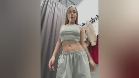 Media: A video of a young Caucasian woman with long blonde hair, wearing a grey crop top and loose shorts, standing in a brightly lit, minimalist dressing room with beige curtains and hanging clothes.