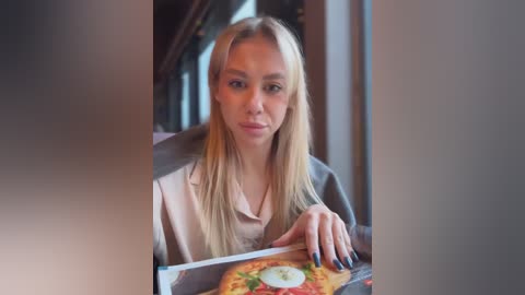 Media: Video of a young blonde woman with straight hair, light skin, and a neutral expression, sitting in a dimly lit restaurant. She wears a beige blouse and holds a pizza with a sunny-side-up egg on it.