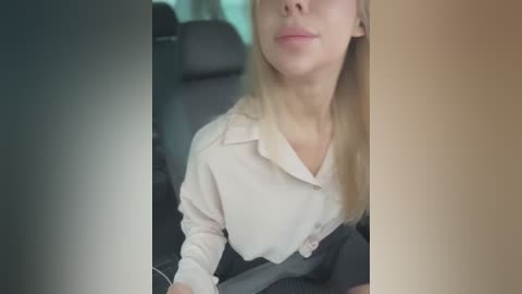 Media: Video of a blonde woman in a beige blouse driving a car. Her face is partially obscured by a vertical split in the image, revealing a calm expression.