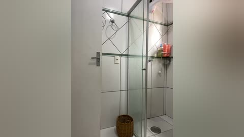 Media: A video of a modern, white-tiled bathroom with a glass shower enclosure, a wicker basket, and shelves holding toiletries.