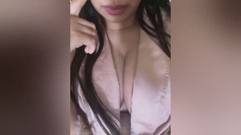 Media: Video of a woman with long dark hair, wearing a low-cut white top, exposing ample cleavage. Her hand is playfully touching her lips. Background is blurred, focusing attention on her.