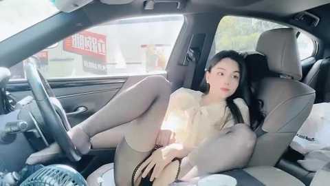 Media: Video of an Asian woman with long black hair, wearing a beige top, black shorts, and stockings, lying provocatively on a car seat. Background shows a car interior and outside store signs.