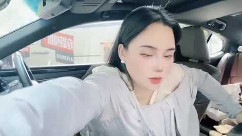 Media: Video of an Asian woman with fair skin and long black hair, wearing a light grey jacket, taking a selfie from a car. Background shows a blurry road sign.