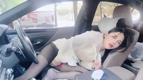 Media: A video of an Asian woman with long black hair, wearing a white blouse, black tights, and a red skirt, lying seductively on a car seat, with blurred background of a street outside.