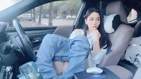 Media: Video of an East Asian woman with long black hair, wearing denim overalls and a white blouse, sitting in a modern car with beige leather seats, looking contemplative.