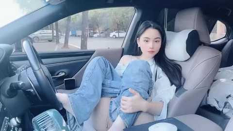 Media: Video of an East Asian woman with long black hair, wearing blue jeans and a white shirt, sitting in a modern car, with a park outside the window.