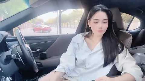 Media: Video of a young East Asian woman with long black hair, wearing a light blue shirt, sitting in a car. Background shows a red car parked outside.